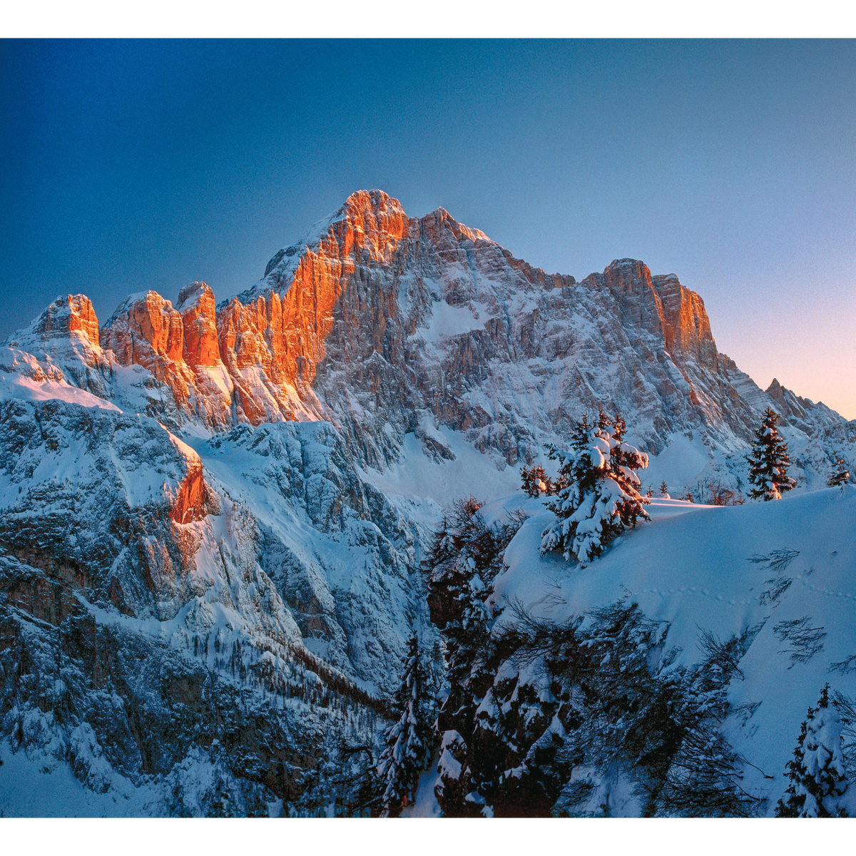 Monte Civetta innevato nelle Dolomiti, con le pareti rocciose illuminate dalla luce arancione del tramonto e abeti ricoperti di neve in primo piano.