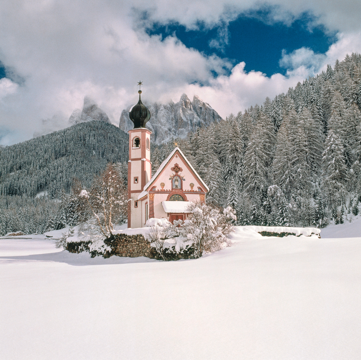 Piccola chiesetta alpina con campanile immersa in un paesaggio innevato, circondata da abeti coperti di neve e montagne sotto un cielo blu e nuvoloso.