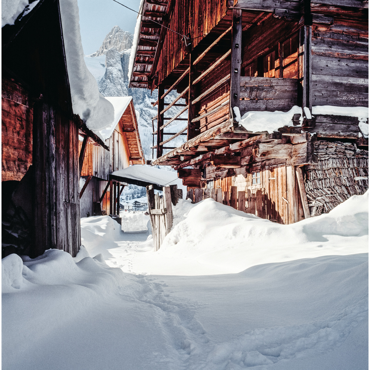 Vicolo innevato in un antico borgo alpino, circondato da case tradizionali in legno e pietra, con la luce del sole che illumina la neve e le montagne sullo sfondo.