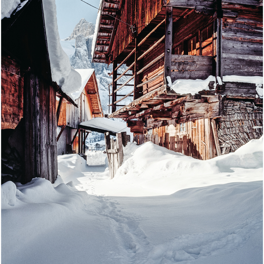 Vicolo innevato in un antico borgo alpino, circondato da case tradizionali in legno e pietra, con la luce del sole che illumina la neve e le montagne sullo sfondo.